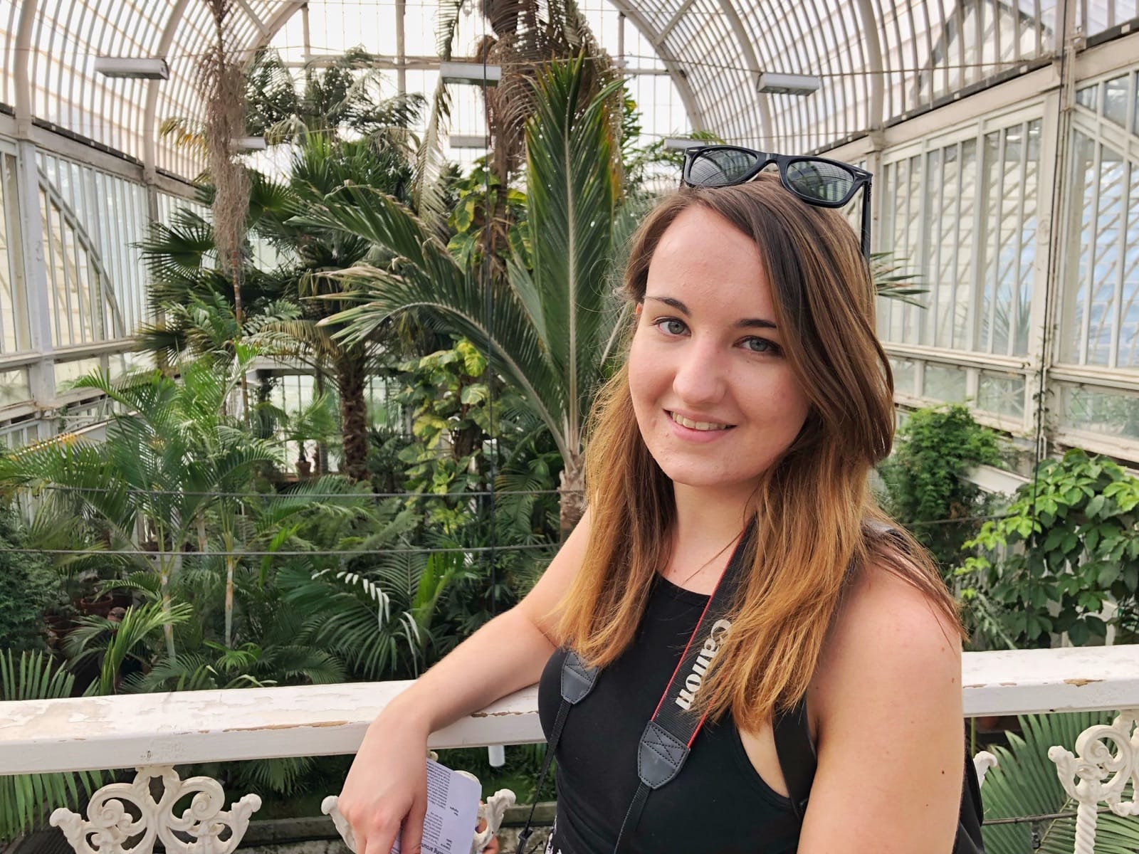 A woman with brown hair leaning on a rail inside of a greenhouse. She has glasses on top of her head.