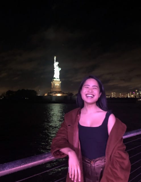 A woman with short dark hair, laughing. The setting is dark, at night time, and the Statue of Liberty is lit up in the distance behind her.