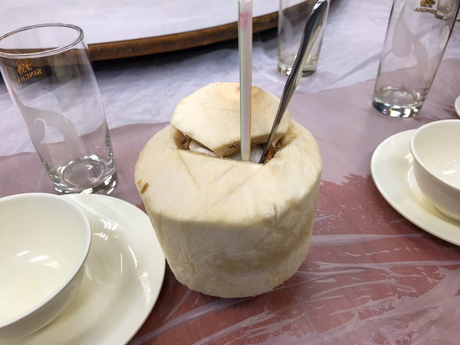 A primitive dining table setting, with a coconut with the top sliced open. It has a straw and spoon sticking out of the top if it.