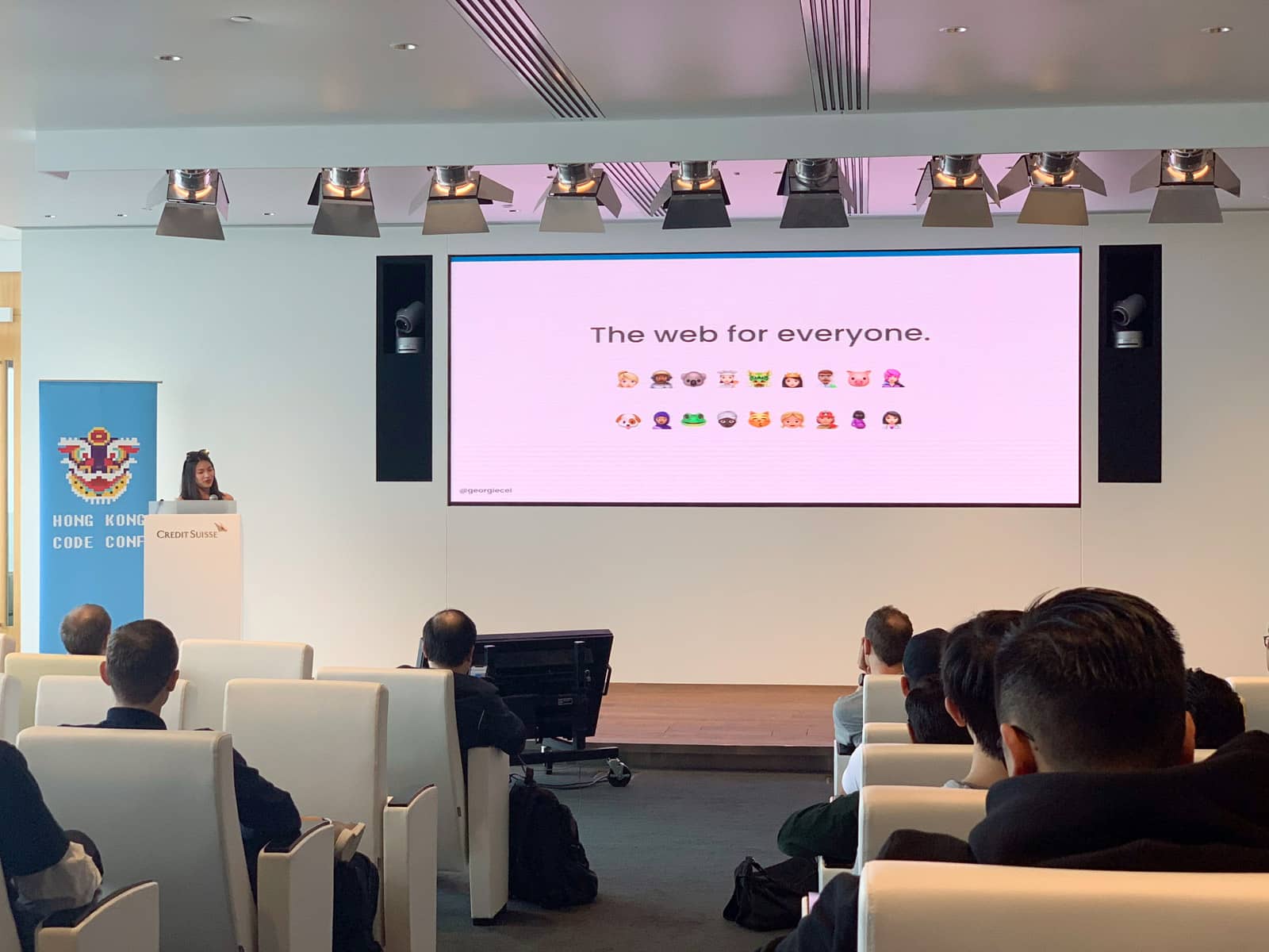 The inside of a conference room with a wide screen showing “The web for everyone” and a number of different emoji to represent diversity. A woman stands at a podium at the front of the room on a stage.