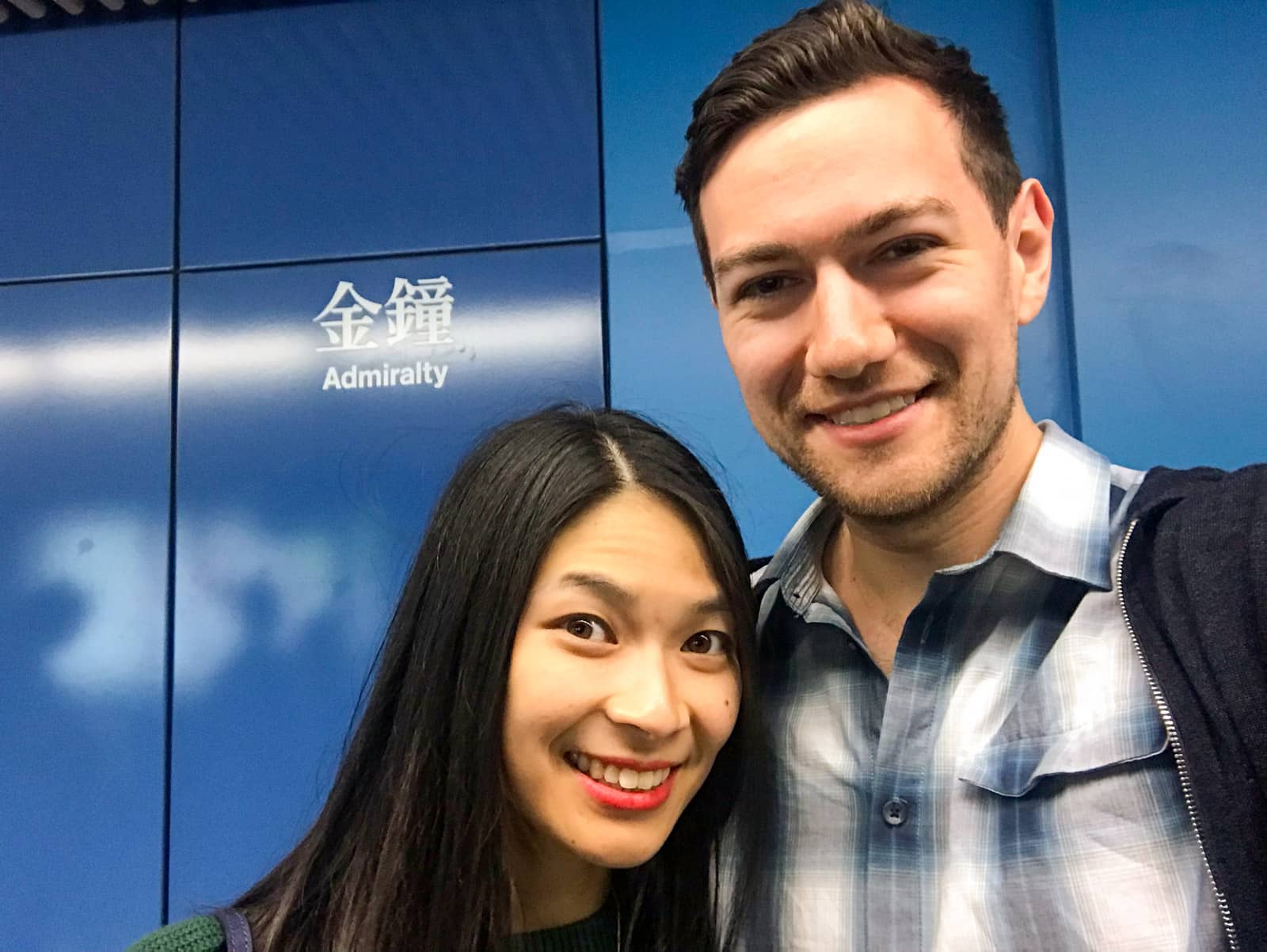 A man and woman with dark hair, smiling. They are taking a selfie in front of a blue wall with “Admiralty” written in English and in Chinese symbols.