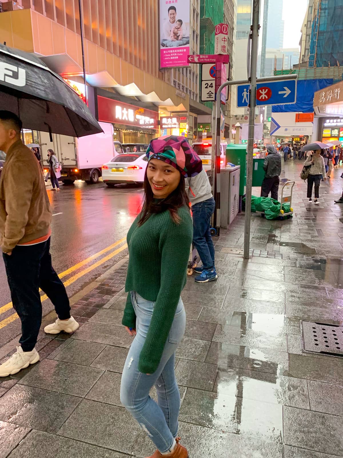 A woman wearing a dark green jumper and light blue jeans and brown boots, with a brightly coloured showercap on her head. She is outside in the rain in a street in Hong Kong