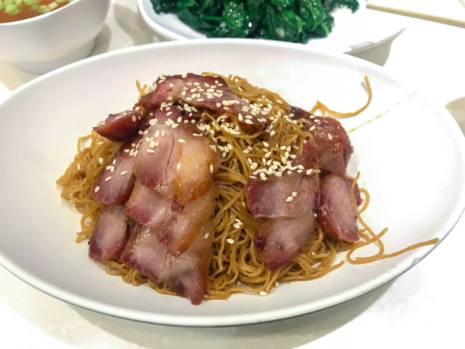 A white bowl of stir-fried noodles served with slices of roast pork, topped with sesame seeds.