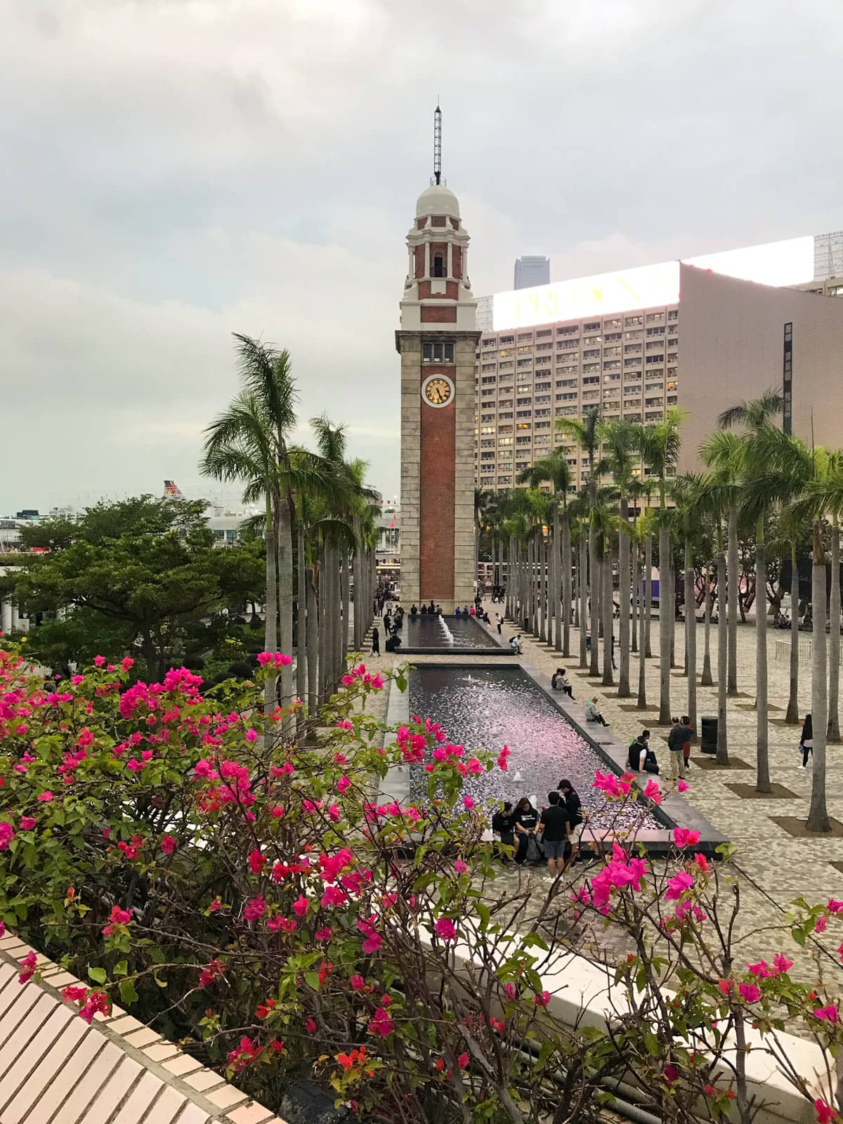 A tall tower with a long water feature in front of it. Palm trees adorn the sides of the water feature. The sky is cloudy and muted in colour.