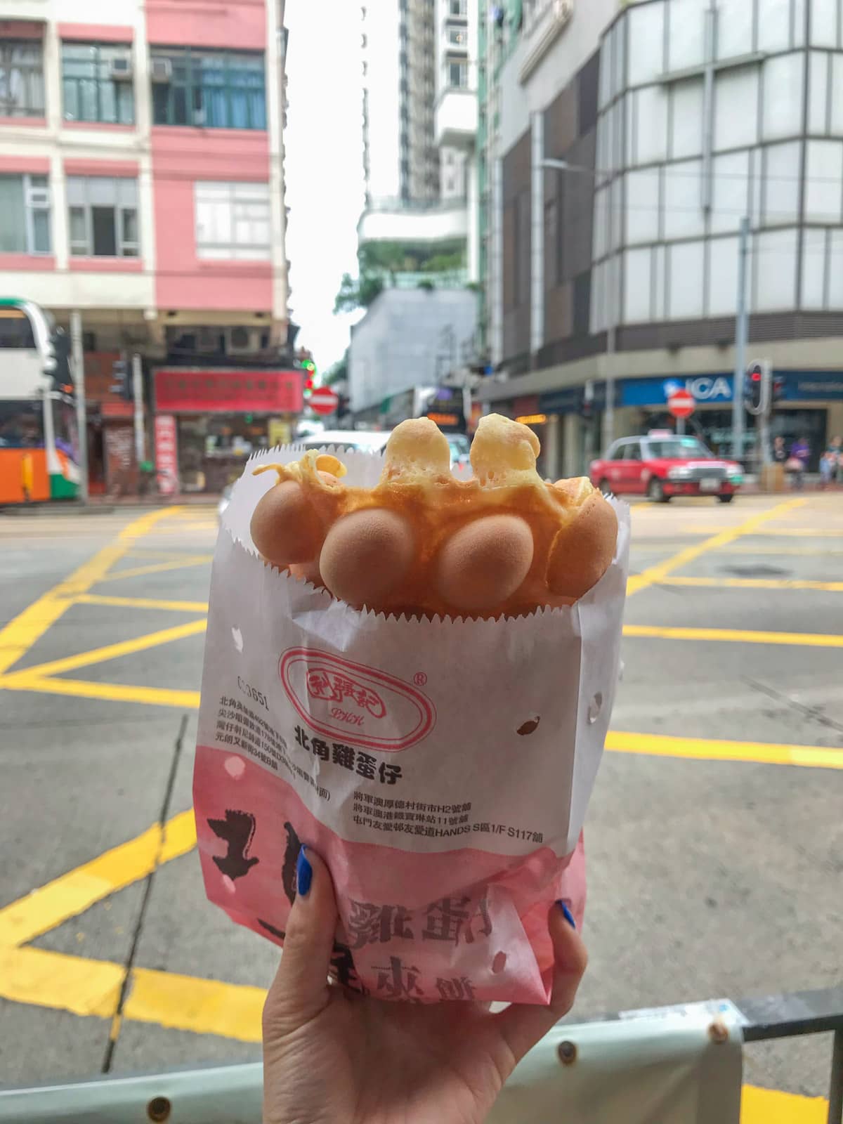 A woman’s hand holding a paper bag with a Hong Kong style egg waffle in it, having the texture of hemispheres