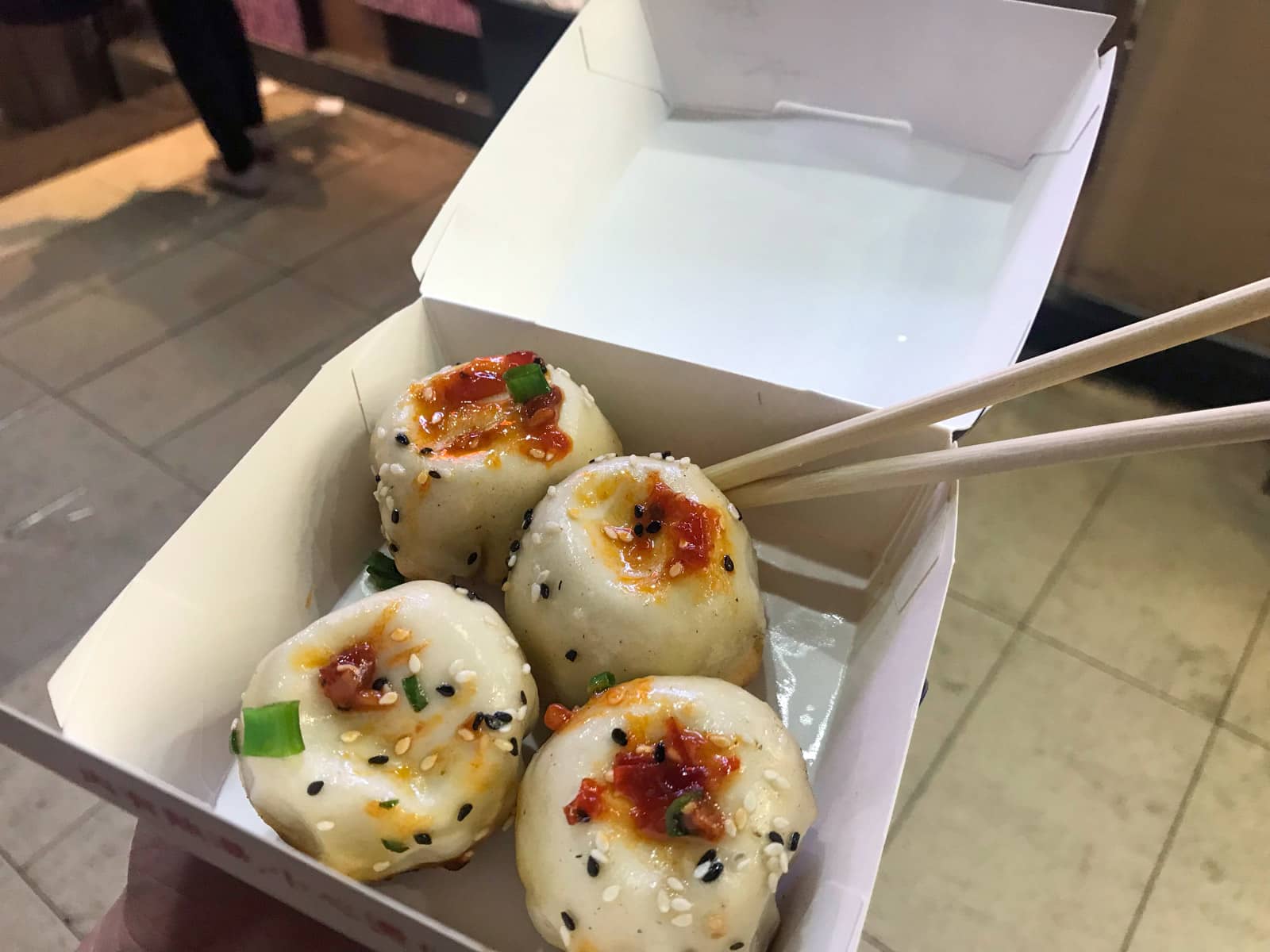 A cardboard box of four pan-fried pork dumplings seasoned with sauce. Wooden disposable chopsticks stick out the side of the box