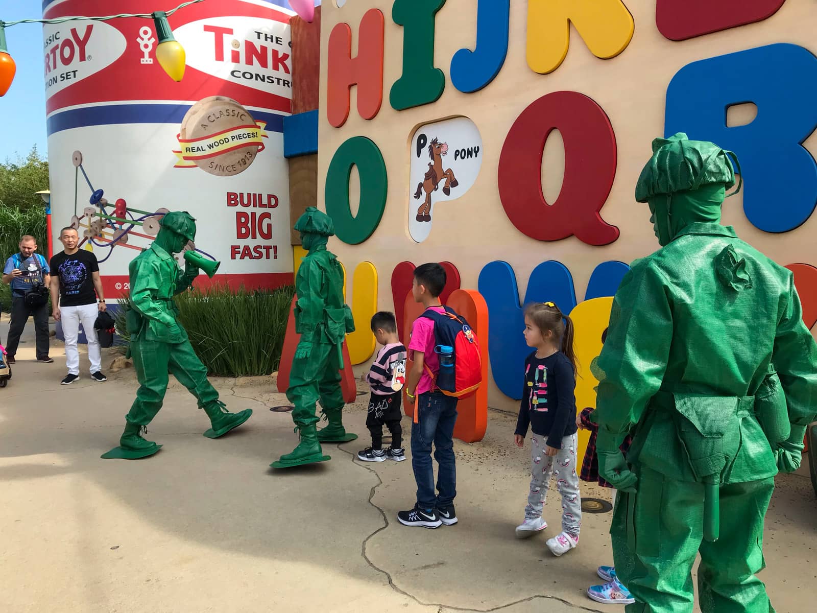 The inside of a theme park with some people dressed in character as life-sized green soldier figurines. A wall with colourful letters of the alphabet sit on the side.