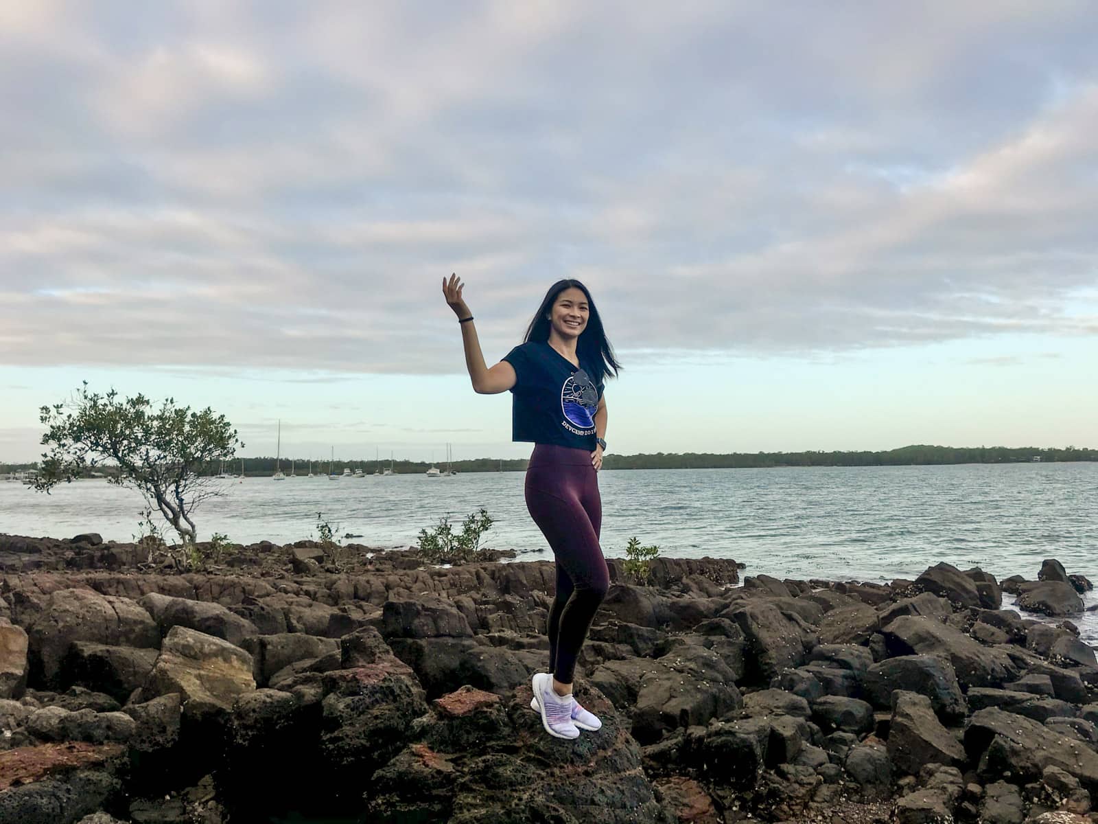A woman wearing dark red leggings, a cropped tee and white shoes, standing on rocks by the sea. One of her hands is up in the air with her palm towards the sky, her other hand on her hip.