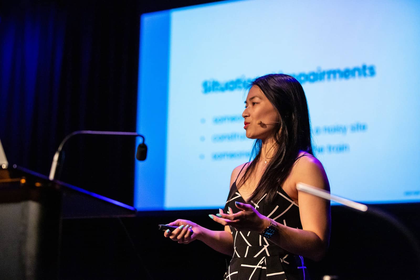 A woman with long dark hair wearing a black and white patterned jumpsuit, standing at a podium delivering a talk. She has a microphone headset.