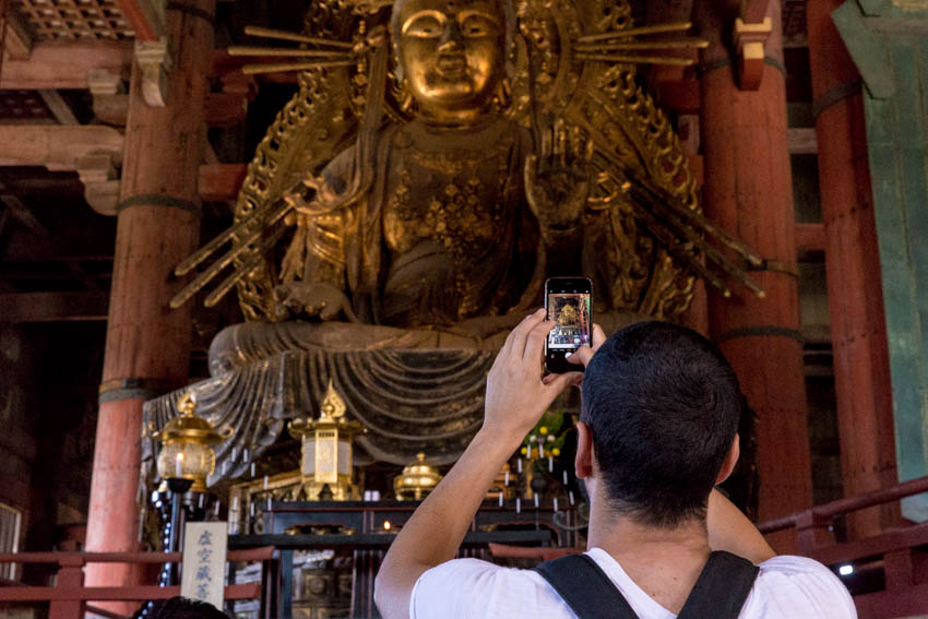 A man in the foreground holding his phone up to take a photo of the bronze buddha in the background