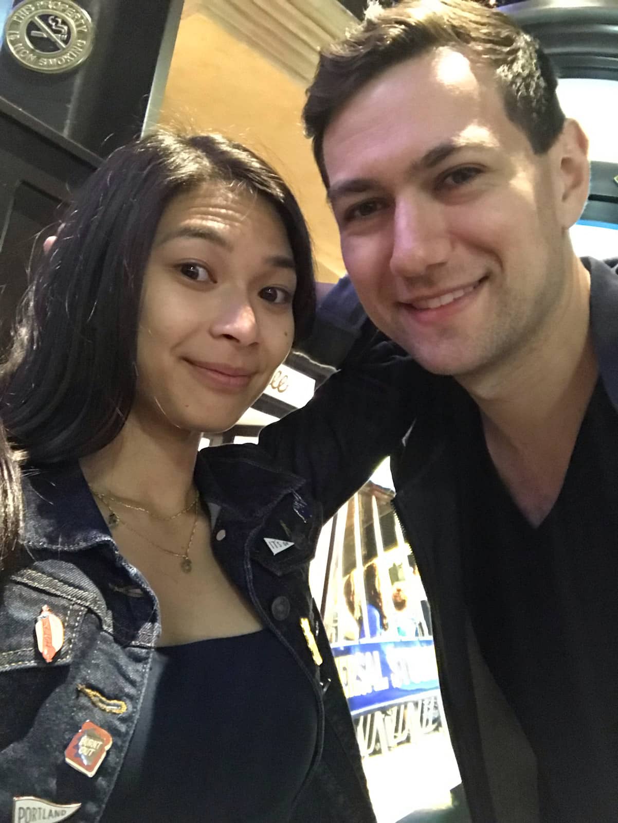 A man and woman taking a selfie and smiling. The woman is wearing a denim jacket with enamel pins and the man is wearing a dark v-neck shirt. The photo is taken in low lighting.