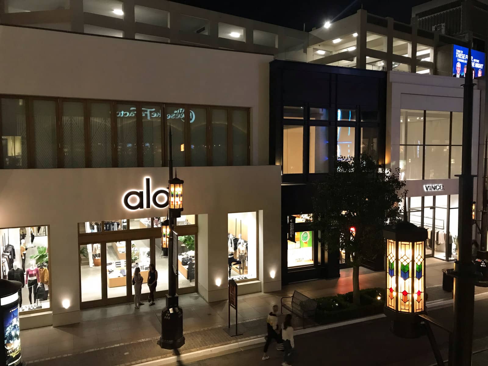 A open-air shopping centre from a balcony, at nighttime. Lampposts and signage provide lighting to the stores, most of which look like they are outside of trading hours.