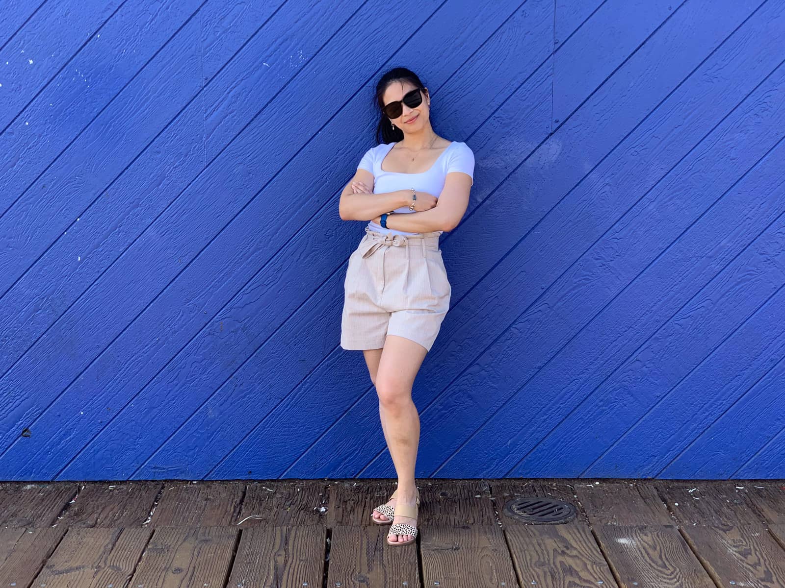 A woman in a white shirt and light coloured shorts, with her arms folded, standing against a wooden backdrop painted in royal blue.