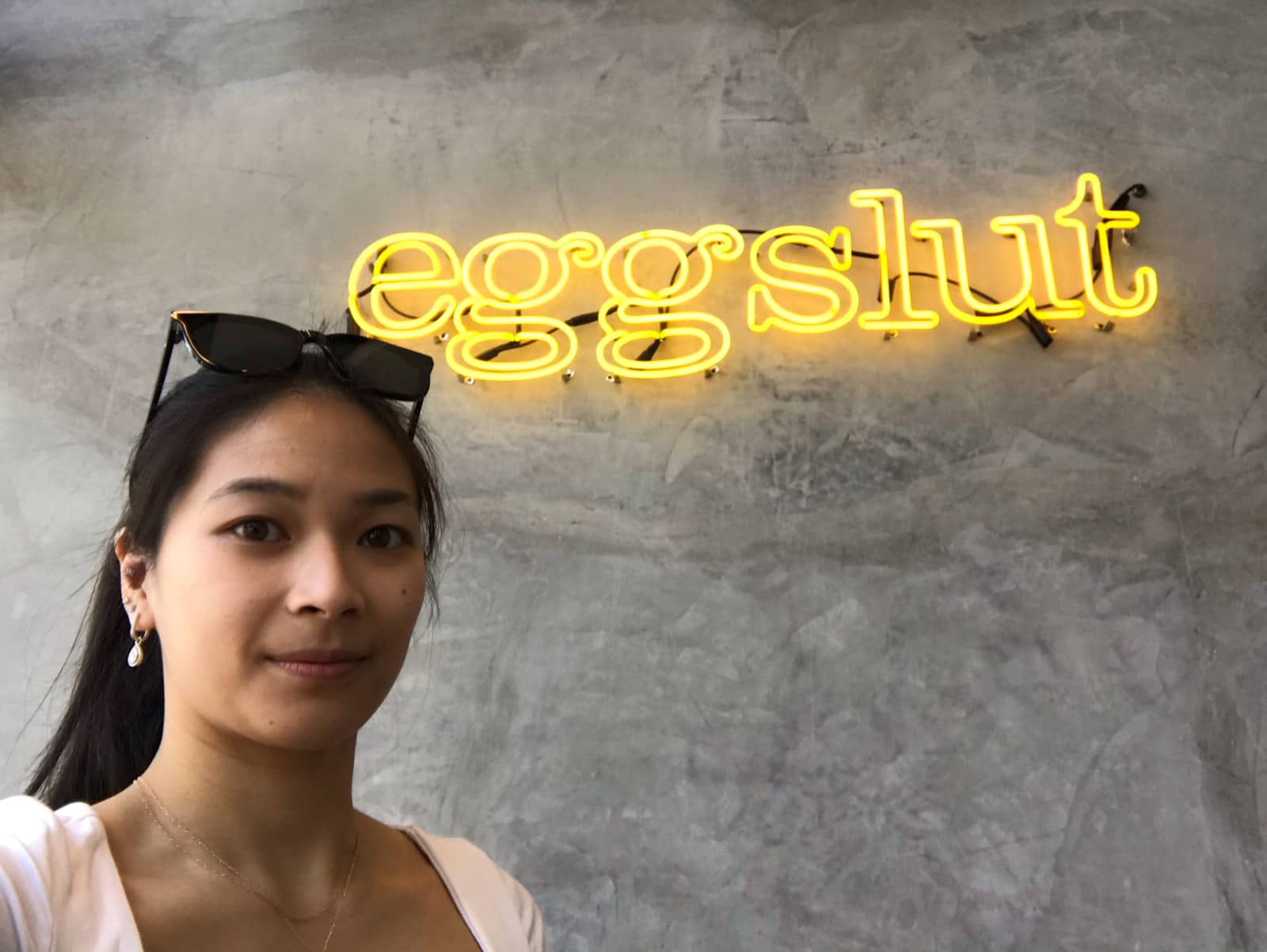 A woman with dark hair wearing sunglasses on top of her head, taking a selfie near a yellow neon sign on a grey wall that reads â€œeggslutâ€�