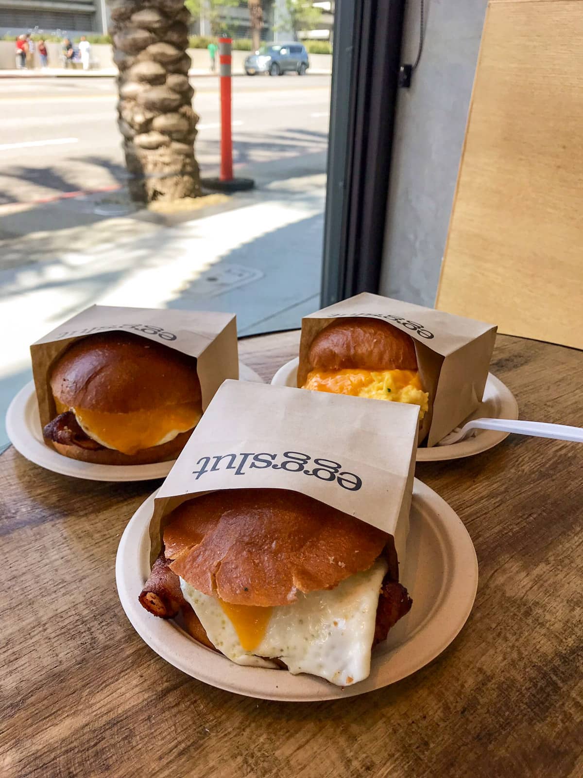Three egg burgers, individually served in small brown paper bags with the text “eggslut” printed on them. They are all sitting on brown paper plates on a wooden table