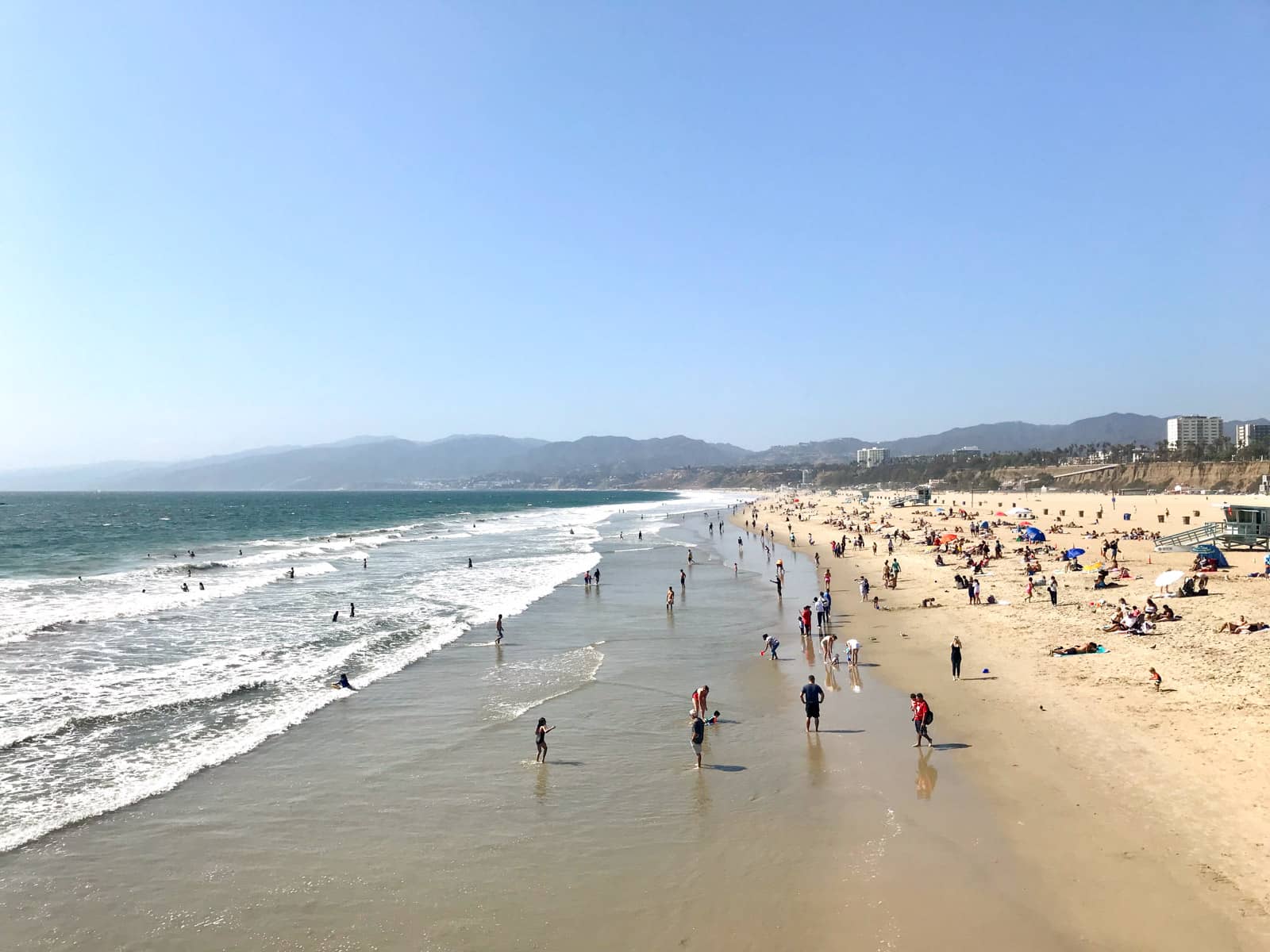 Santa Monica Beach in Los Angeles, seen from the boardwalk. The sea is on the left had side and the sandy beach is on the right. The beach has many people standing in the shallow water and many with umbrellas and sitting on the sand