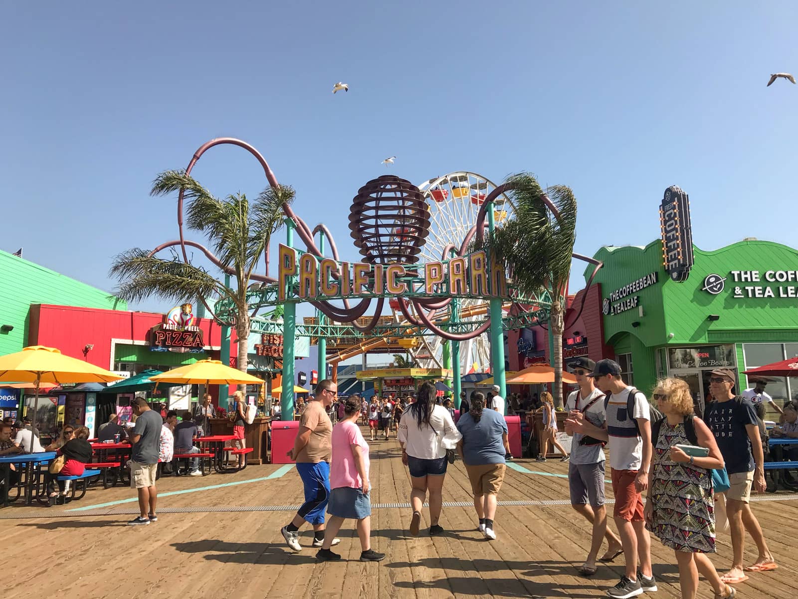 The entrance to an amusement park, called Pacific Park. It is situated on a pier. A pizza parlour sits on the left of the entrance and a coffee and tea shop on the right. The rides inside the park, and the signage, are seen to be brightly coloured