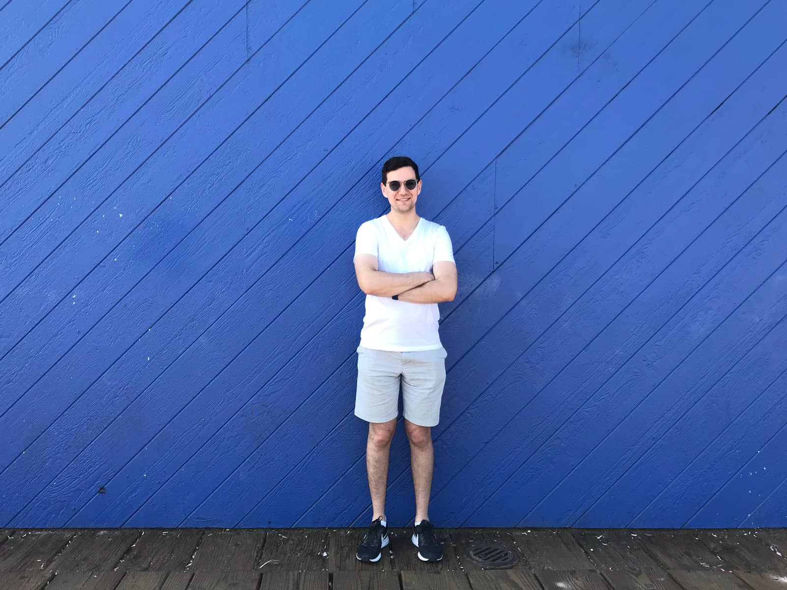 A man in a white shirt and light coloured shorts, with his arms folded, standing against a wooden backdrop painted in royal blue.
