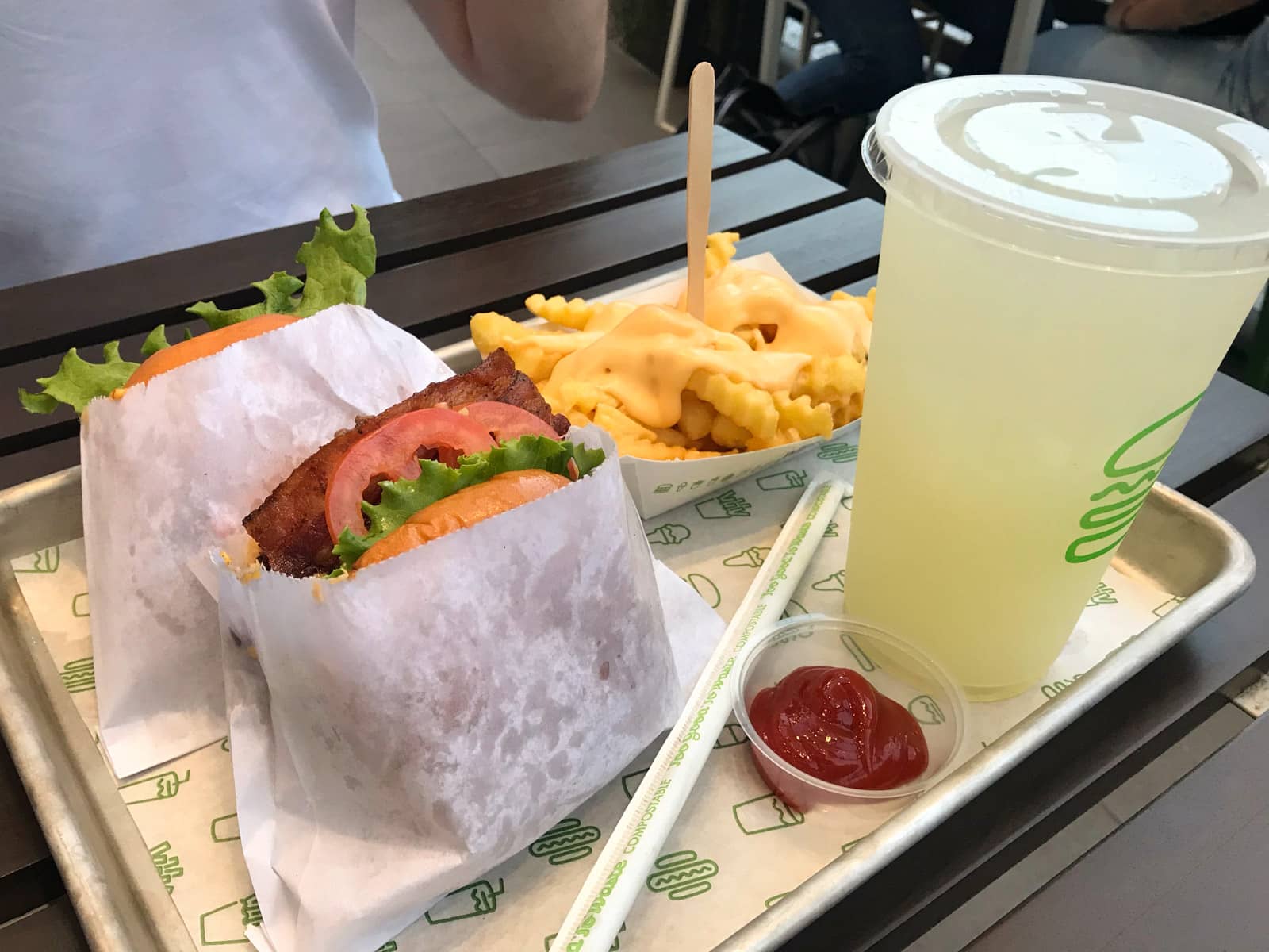 A silver tray served with two burgers which can be seen to be filled with bacon, tomato and lettuce. Also on the tray is a small box of crinkle cut fries, a plastic cup of lemonade, and a small saucer of tomato sauce