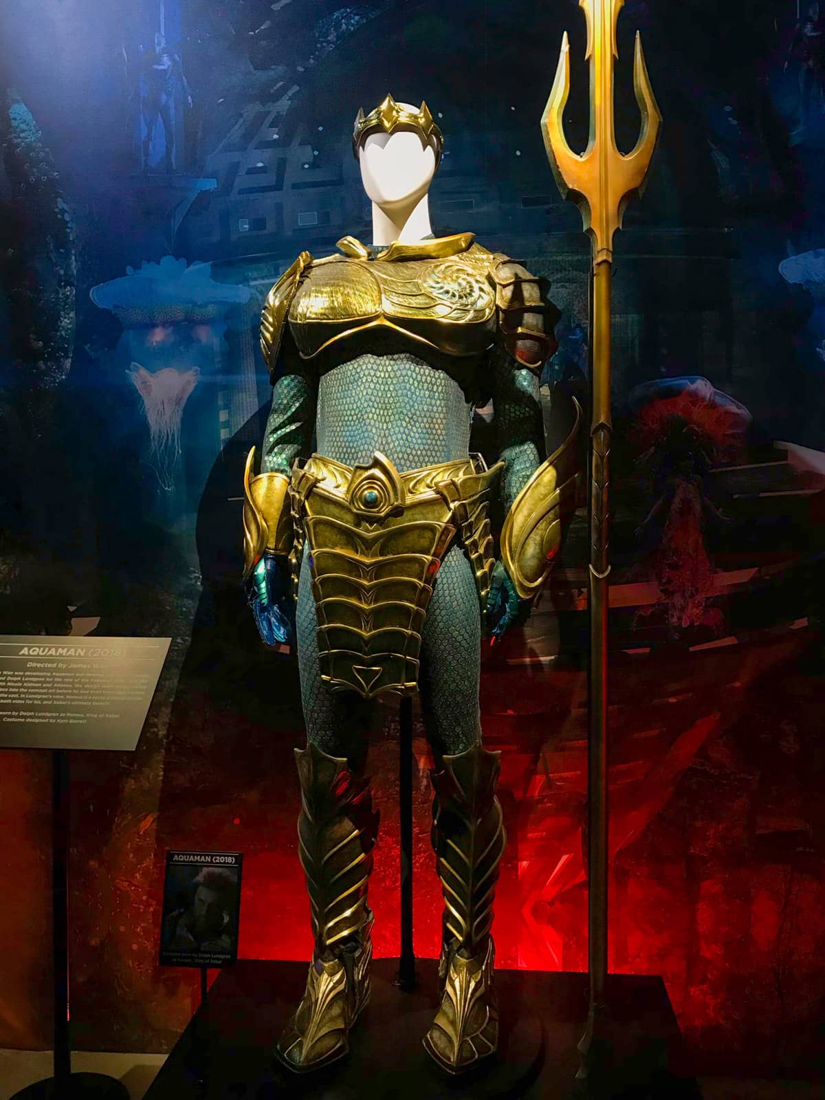 An Aquaman costume on display on a mannequin. The cost is metallic blue and gold with shiny detail.  His trident stands next to him.