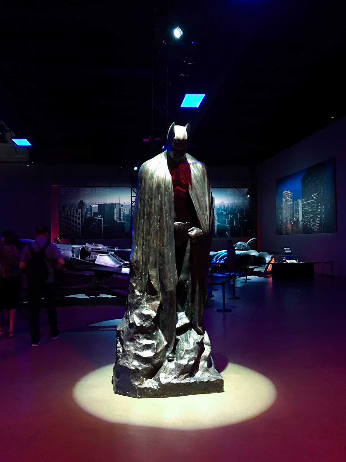 A dark room with a statue of the Batman character, with a spotlight light from above. Batman is looking down towards the ground. In the background are some automobiles