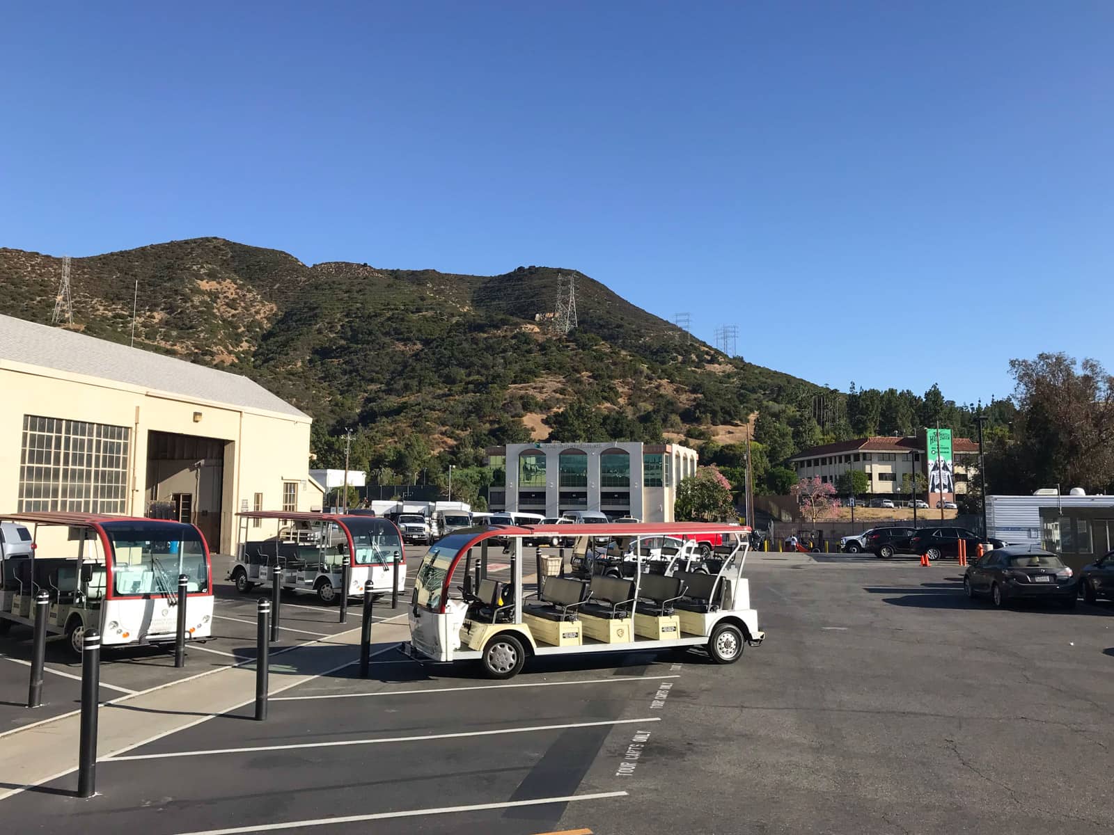 The outside of a film and television studio, with open passenger buses parked outside. In the background you can see the hills of Los Angeles, brown in colour but dotted with many trees.