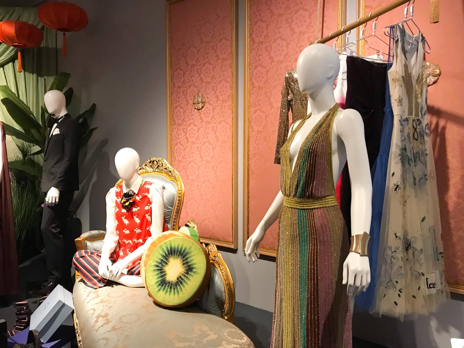 Mannequins on display with a couple of different characters, one sitting on a couch and the other in a ballgown. A rack of clothes stands behind one mannequin. There is a kiwi-fruit-styled cushion on the couch.