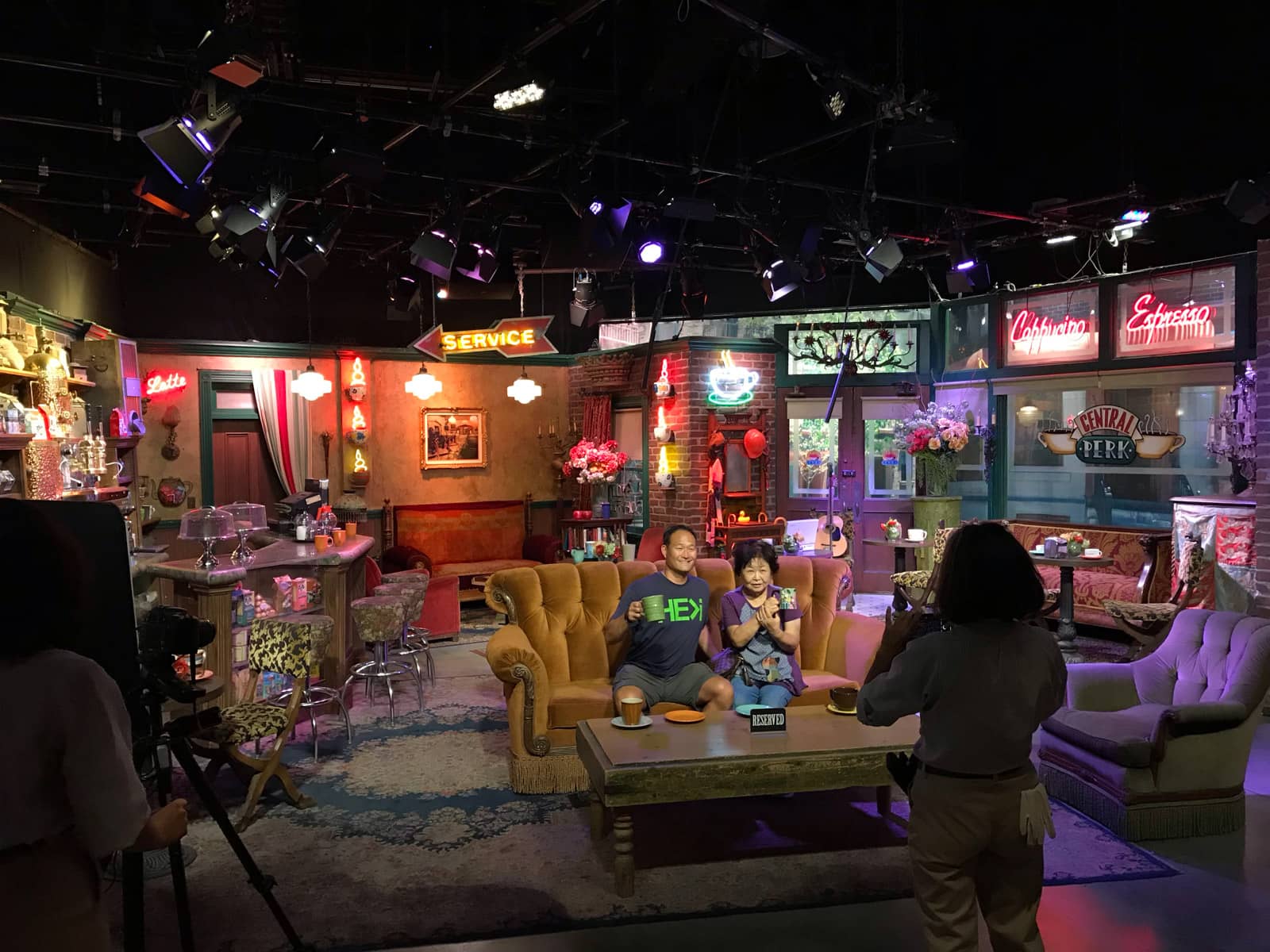 A television stage set with various props and neon signs. In the centre of frame is a large old couch with two people sitting in it, posed for a photo