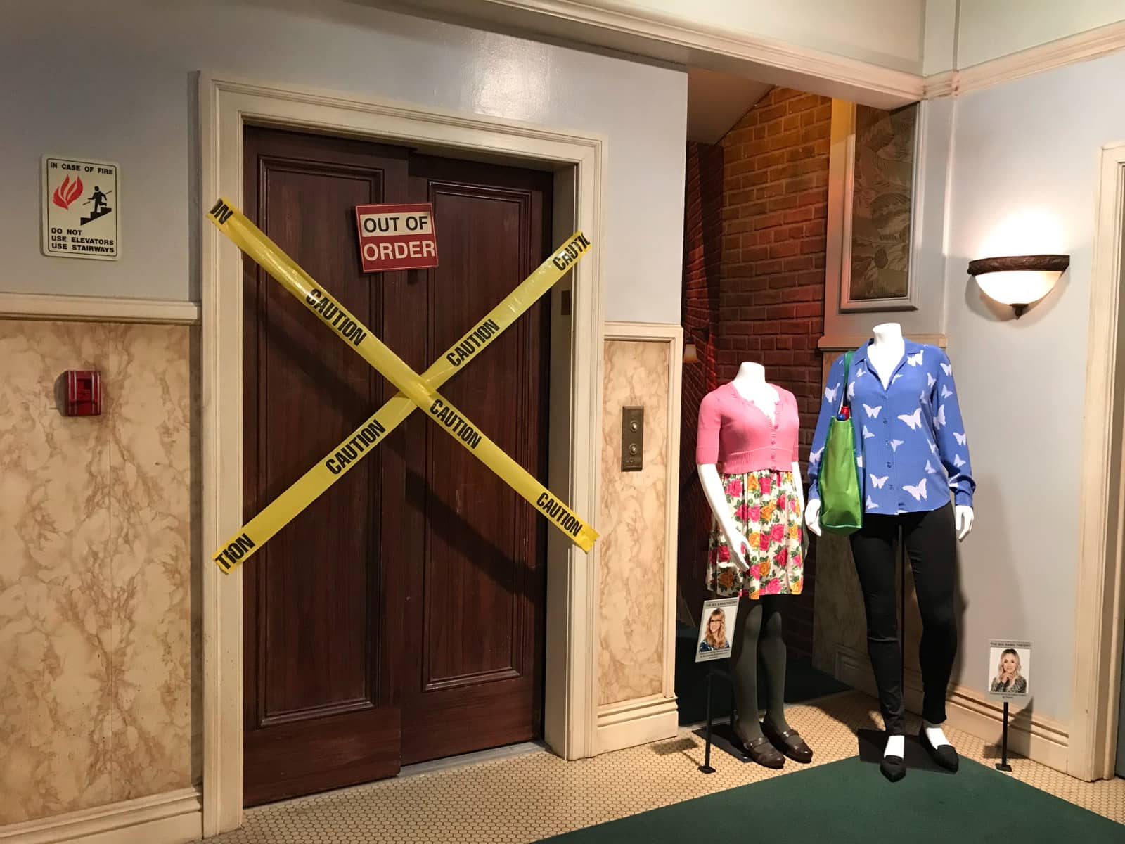 A television stage set that looks like the elevator in an apartment block, but that is not functioning. Yellow tape blocks the doors of the elevator. Two mannequins stand next to the elevator door, displaying characters’ costumes.