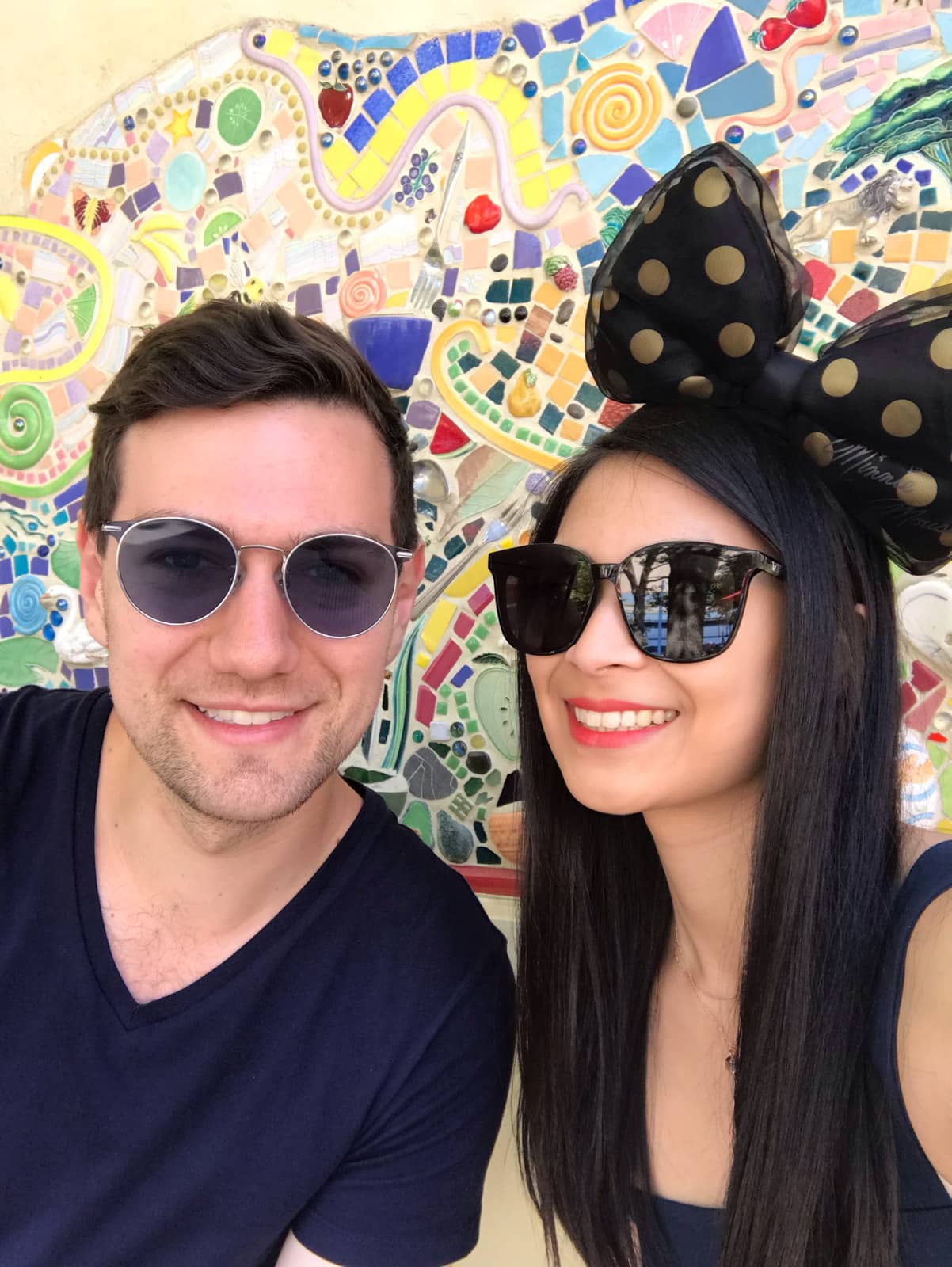 A man and woman in a selfie, in front of a colourful mosaic tile wall. They are both smiling and both wearing sunglasses. Both of them have dark hair. The woman is wearing a big bow on top of her head that is black with gold spots.