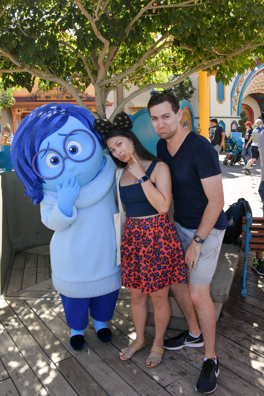 A man and woman posing with sad faces, alongside a person dressed in a character costume of Sadness from Pixar film Inside Out. Sadness’ costume is different shades of blue with blue skin, blue glasses and blue hair.