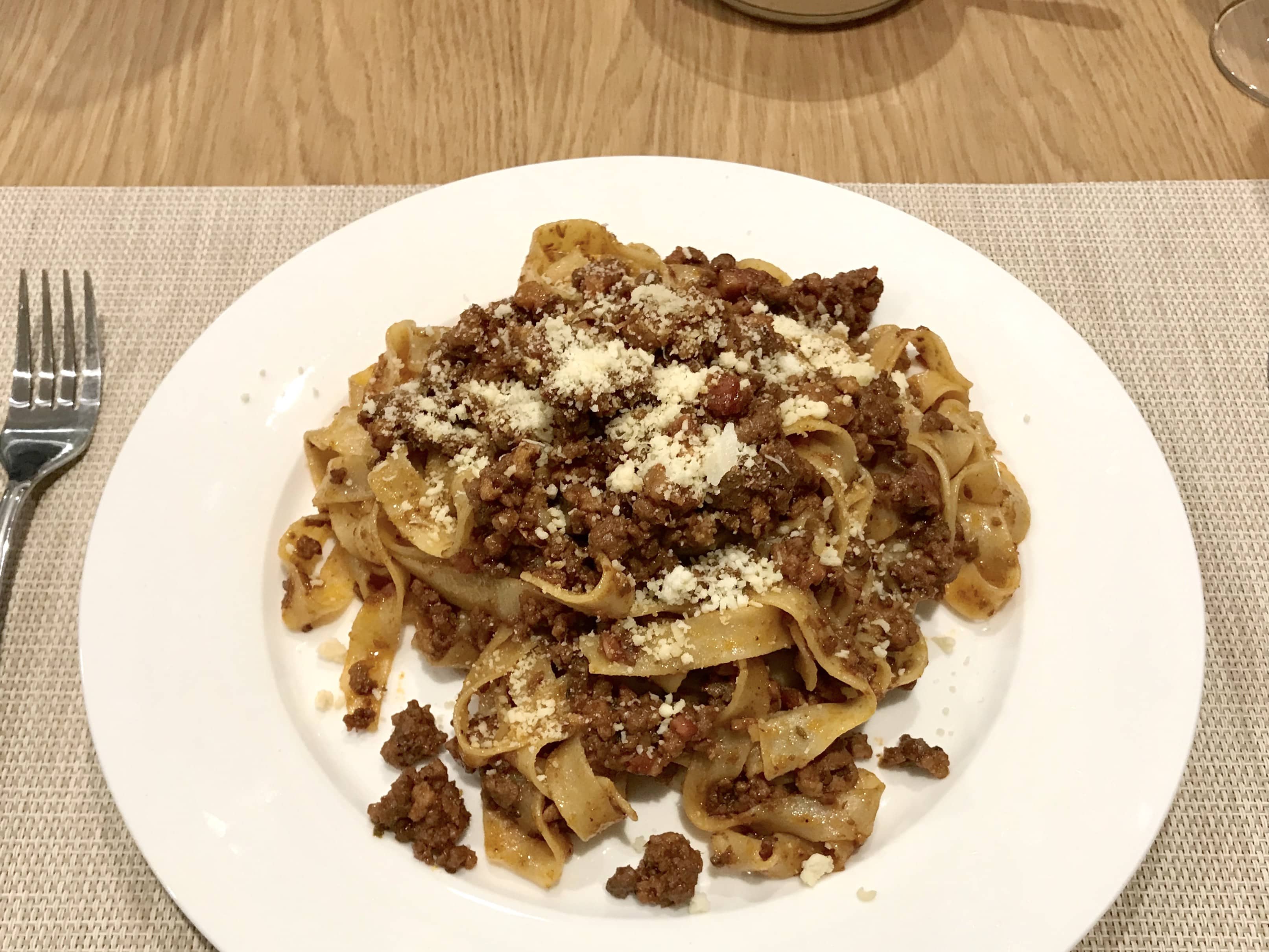 A white plate of spaghetti bolognese, placed on a natural coloured placemat on a brown table.