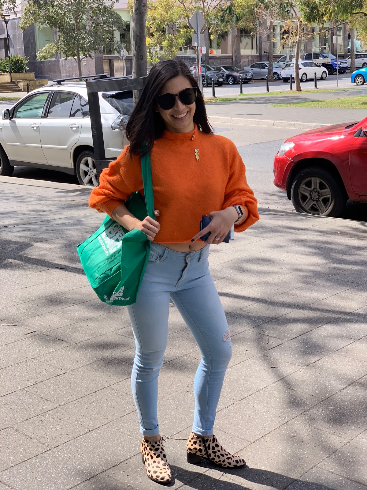 A woman with sunglasses wearing an orange sweater and light blue jeans. She is holding a green grocery bag over her arm.