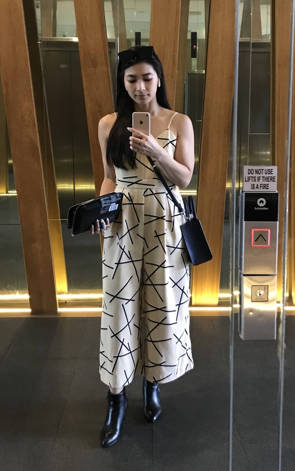 A woman with long dark hair wearing a beige sleeveless wide-leg jumpsuit, taking a selfie in a mirrored surface