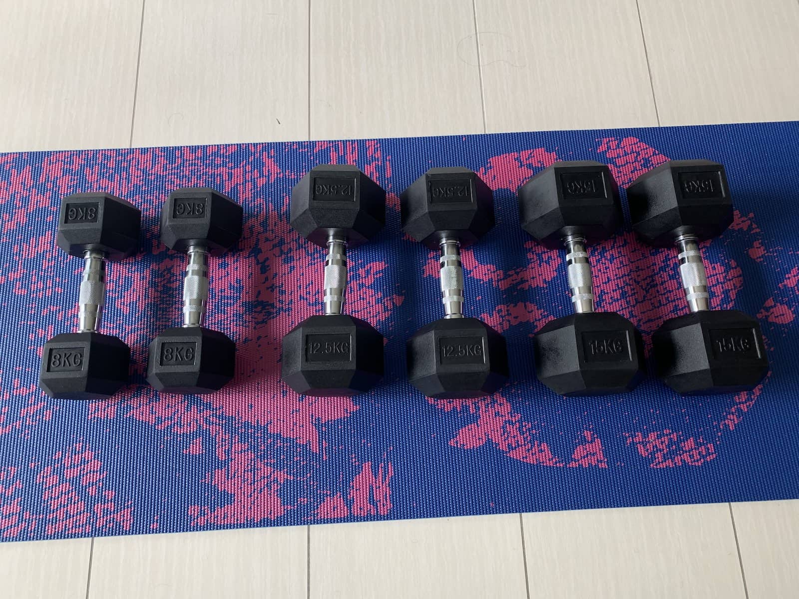 A dark blue yoga mat with a pink pattern. On the mat are three pairs of dumbbells of 8kg, 12.5kg and 15kg.