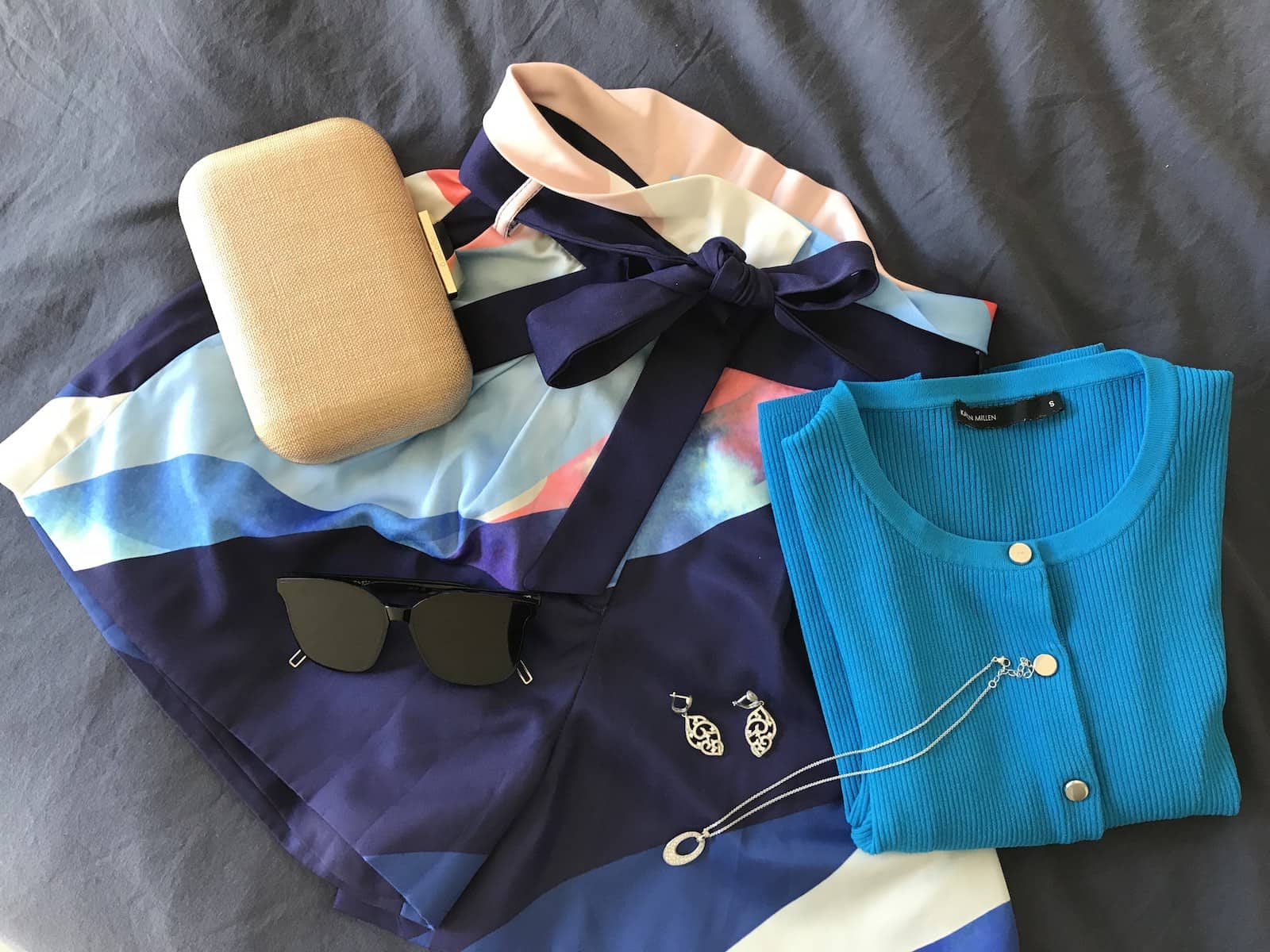 A flatlay of a pair of shorts with white, blue and pink print, with a folded bright blue cardigan, and some jewellery and sunglasses on top.