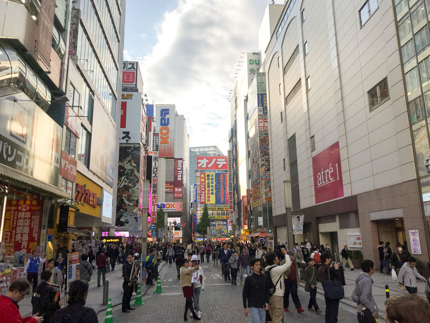 A pedestrian-friendly street between medium-rise buildings. The buildings have colourful signage printed with Japanese katakana and the occasional roman letter. The street is busy with people, some of whom are taking photos