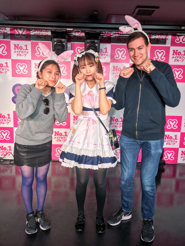 Me, a girl dressed up as a maid, and Nick, with our fists near our face, resembling a cat’s paws. Nick and I are wearing headbands with pink and white rabbit ears on top. Hot pink and white wallpaper with hearts and Japanese writing advertise the café.