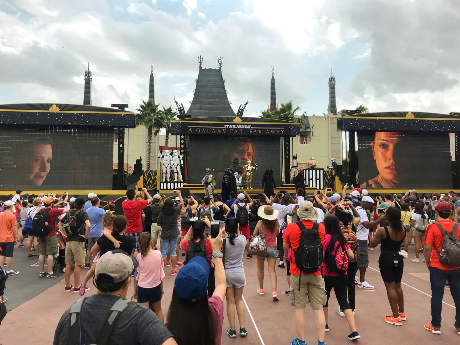 An outdoor stage with screens. There are people dressed up in character. In the foreground are audience members, many recording footage on their smartphones.