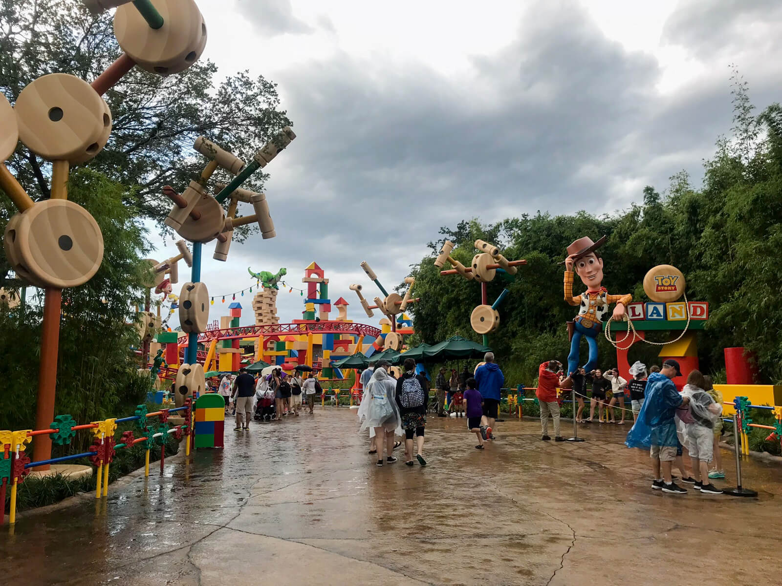 The entrance to an area of a theme park, with statues resembling life-size toys at the edges of the path. All the statues are colourful or resemble wood. The path is very wet and the sky is cloudy.