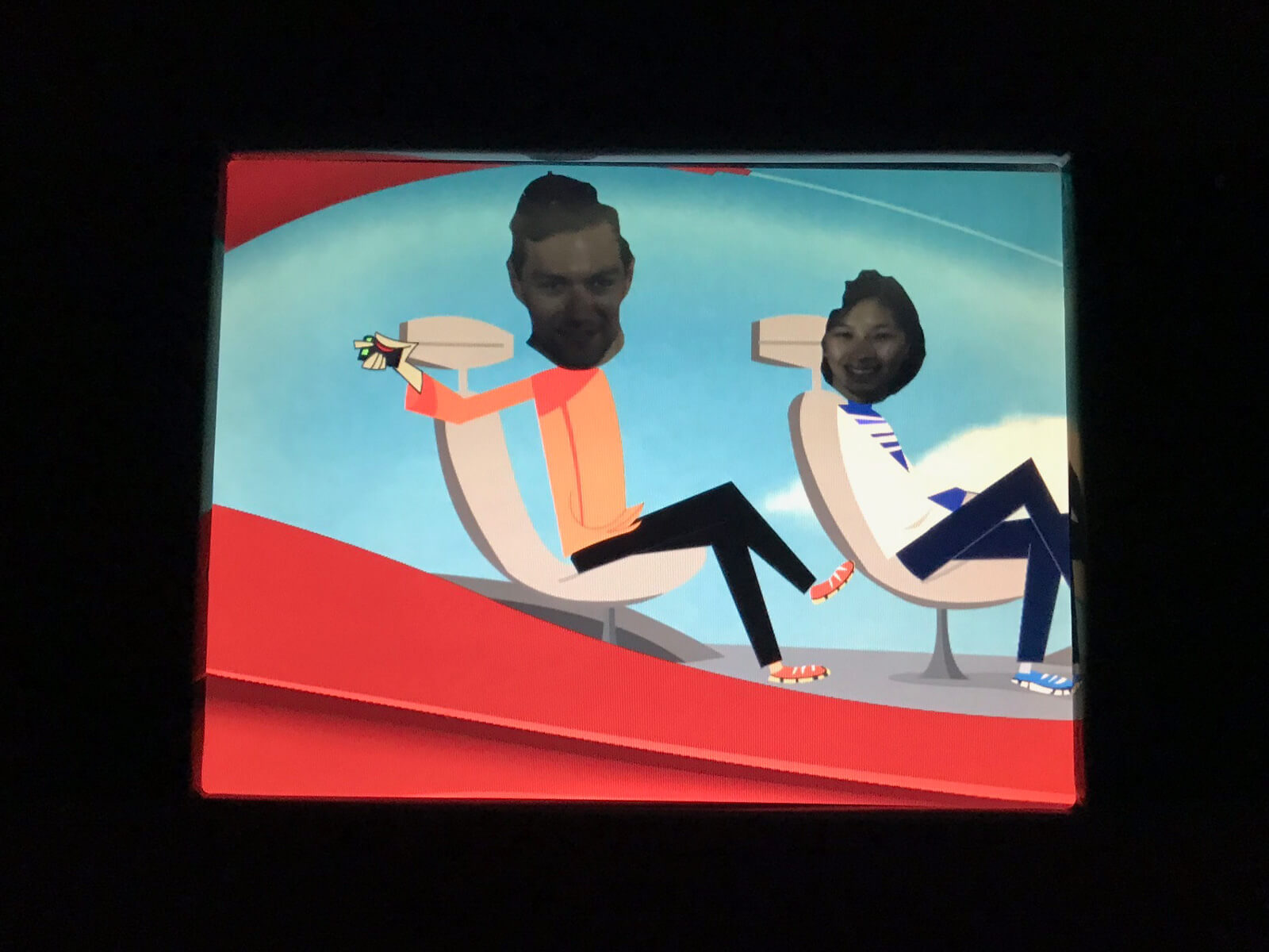 A screen showing two cartoon figures on chairs, with a manâ€™s face and a womanâ€™s face superimposed on them