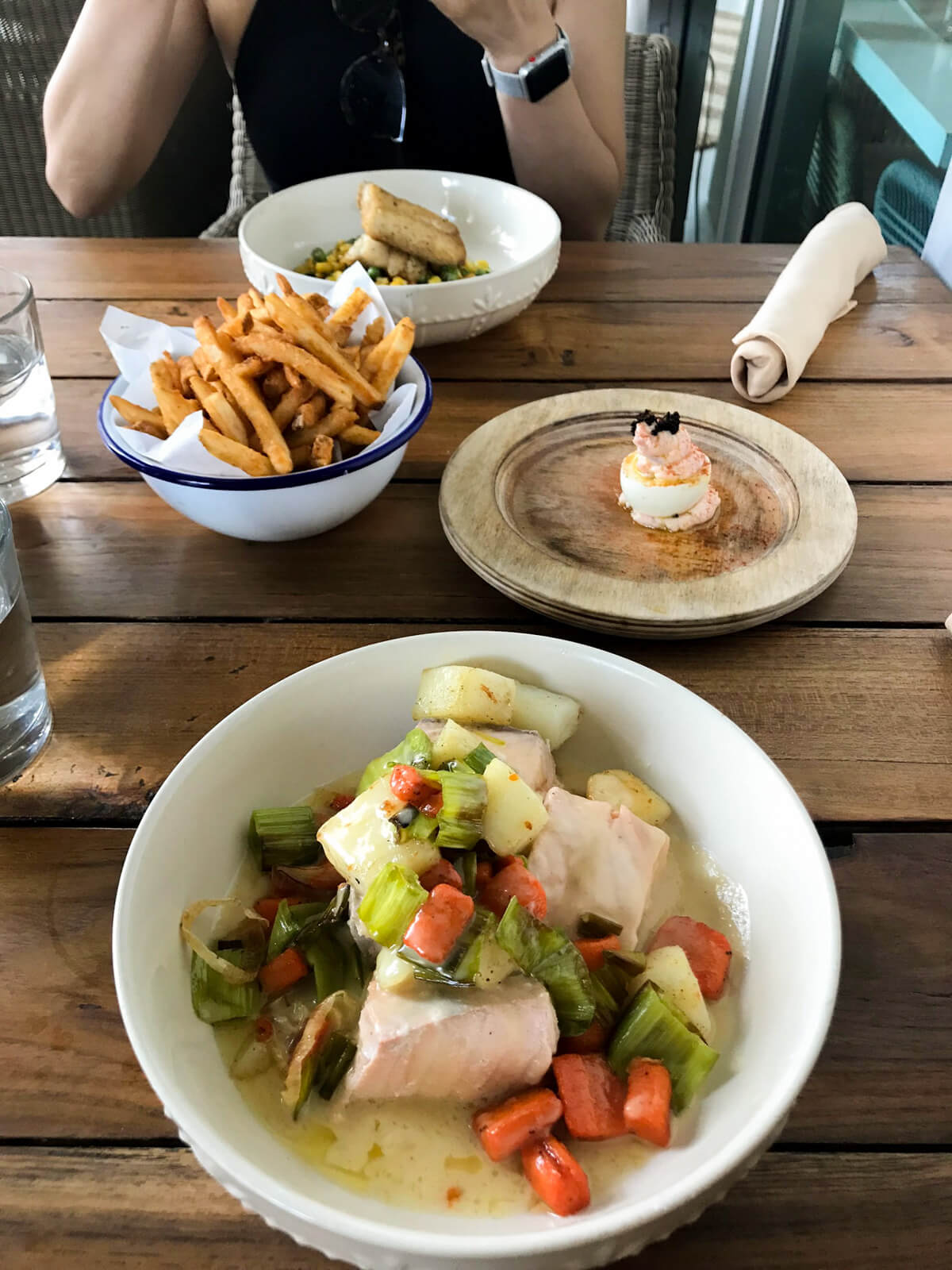 A bowl with cooked fish and vegetables with a soupy base. It’s sitting on a wooden table, with a bowl of chips, another dish consisting of an egg decorated with seafood, and a bowl in the distance also containing fish and vegetables