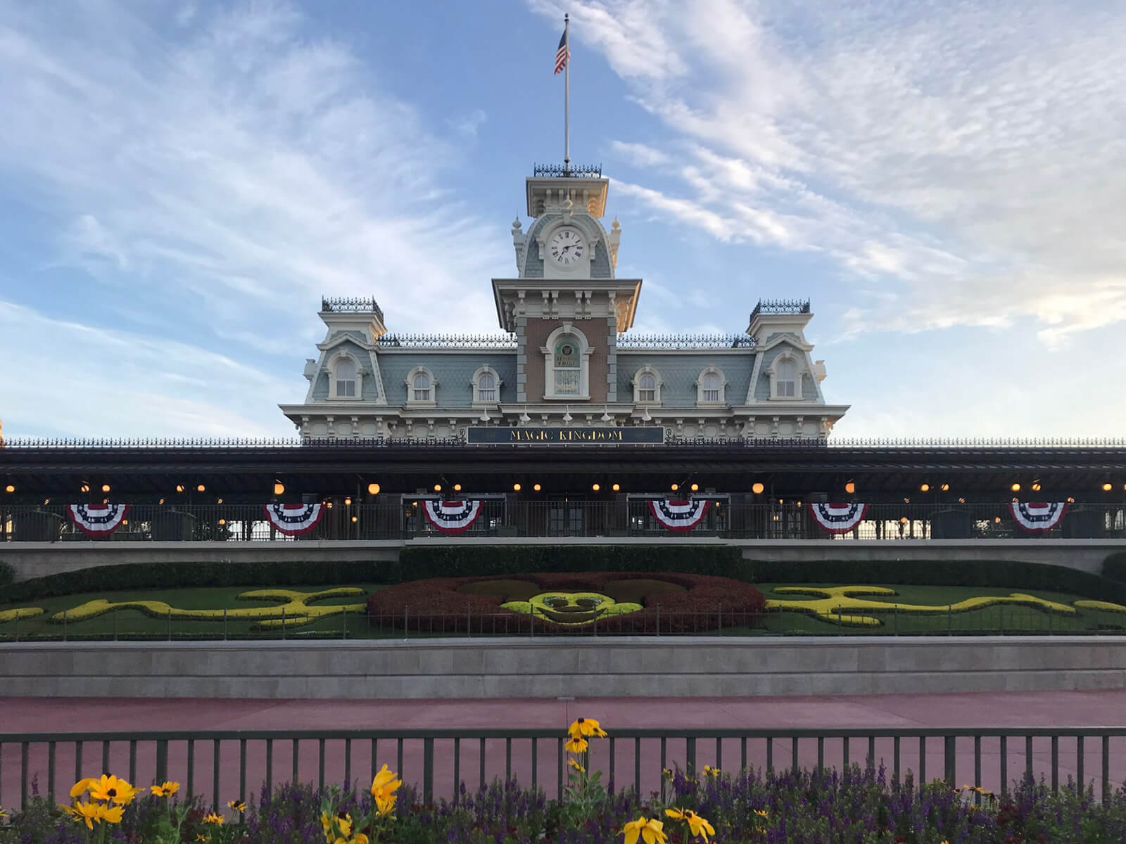 The entrance to Disney Worldâ€™s Magic Kingdom. In the foreground is a garden with a hedge shaped like Mickey Mouse. In the background is a train station with the flag of the United States of America standing from a pole