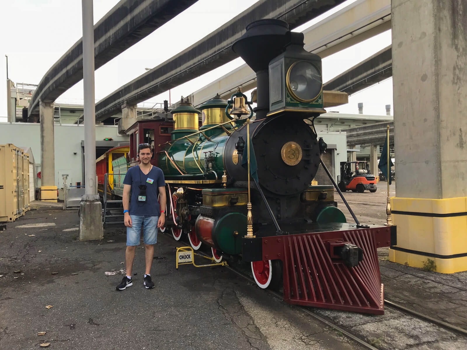 A man with a blue t-shirt and shorts standing next to a green and dark red steam locomotive