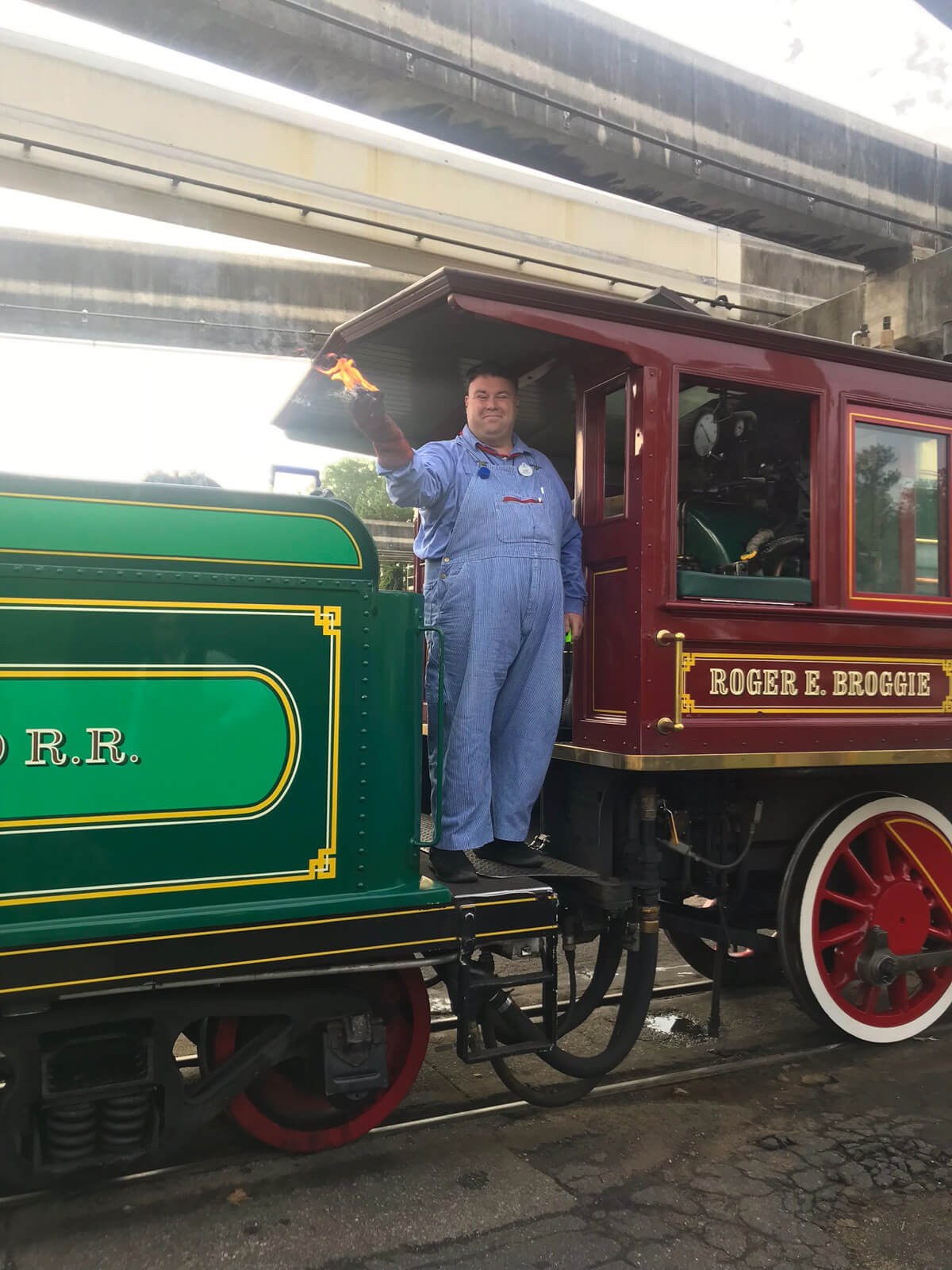 A man in blue overalls holding a piece of coal on fire. He is standing on a steam train