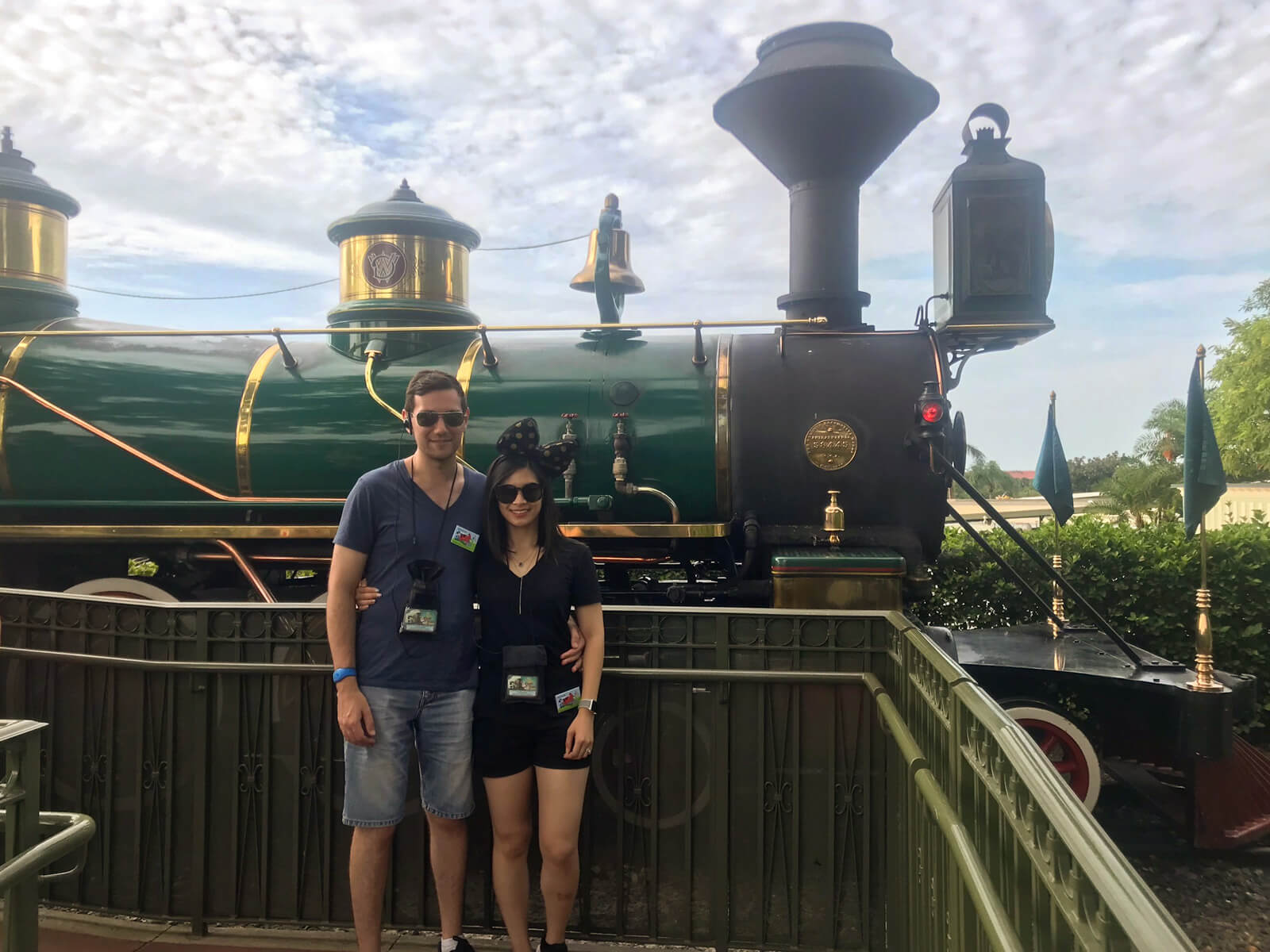 A man and woman standing in front of a green steam train. They are wearing sunglasses and have their arms around each other.