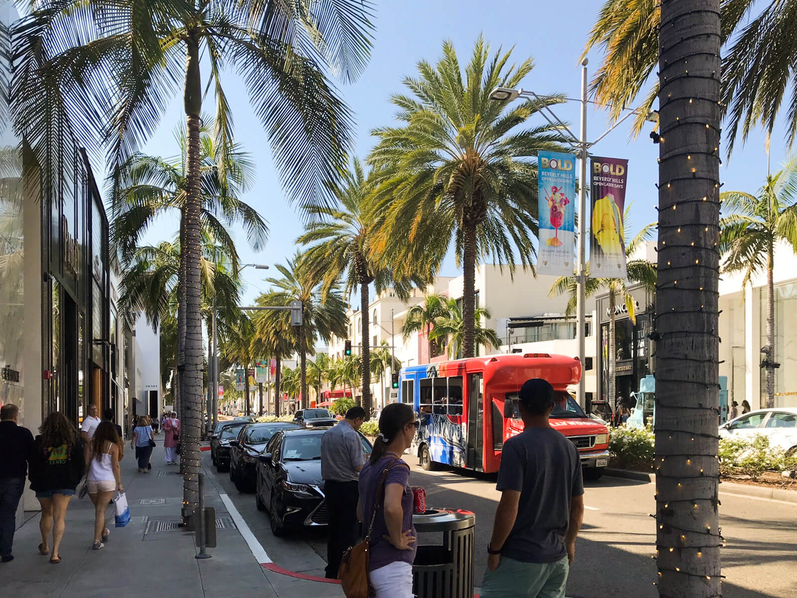 A street with palm trees down the side of the street. The tree trunks have fairy lights wrapped around them. There are cars parked down the side of the street and people walking on the sidewalk.