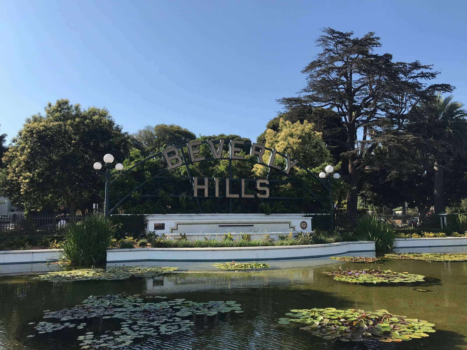 A pond with some lilypads. Behind the pond are trees and a metal sign with the words “Beverly Hills” on it in metal letters