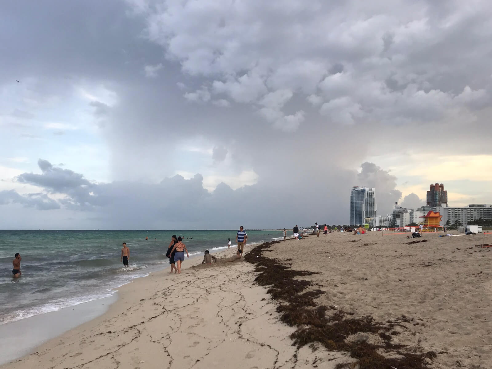 A beach on a very cloudy afternoon, with the sea on the left and some people walking around in swimwear. The sand is light brown and has a long line of seaweed going into the distance along the length of the beach.