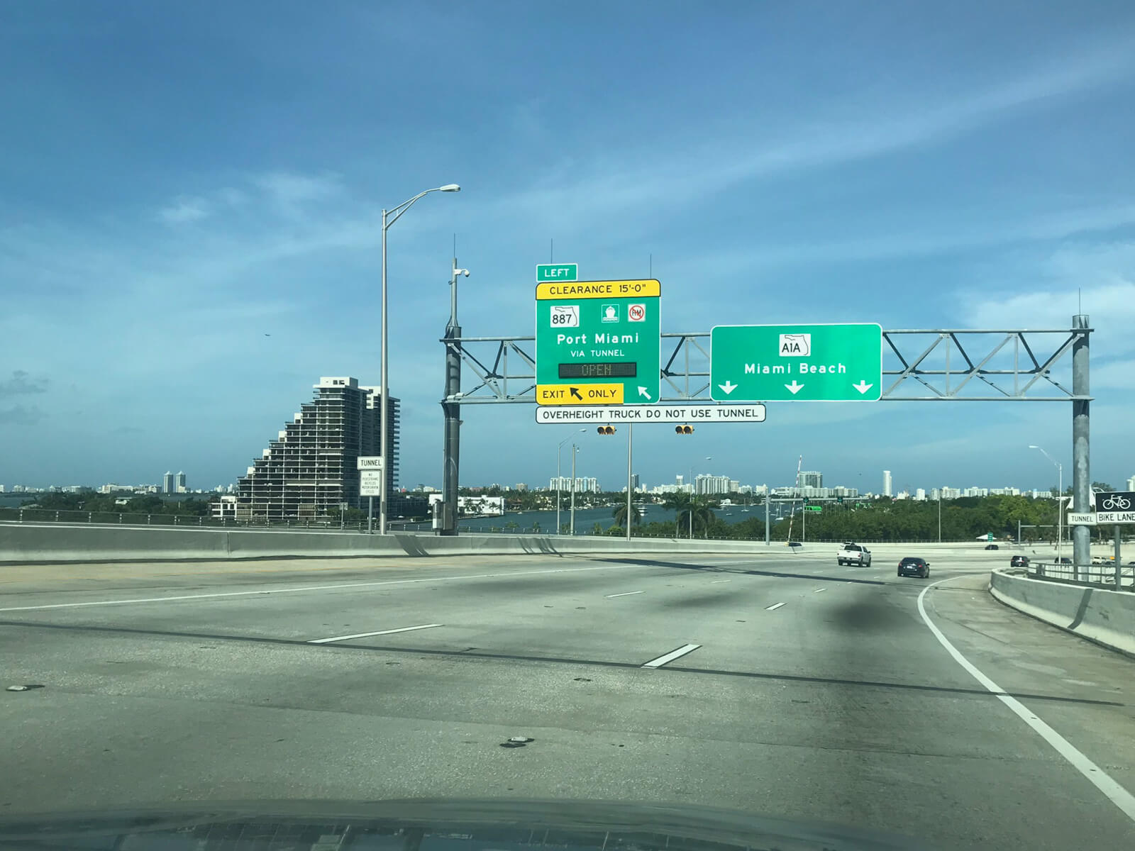 A view of a wide multi-lane elevated highway from the inside of a car. Large green road signs read “Port Miami” and “Miami Beach” and the sea is visible in the distance